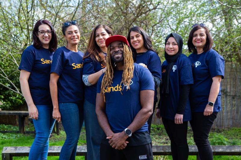 A group of people in Sands charity t-shirts, stood together outside and smiling at the camera.